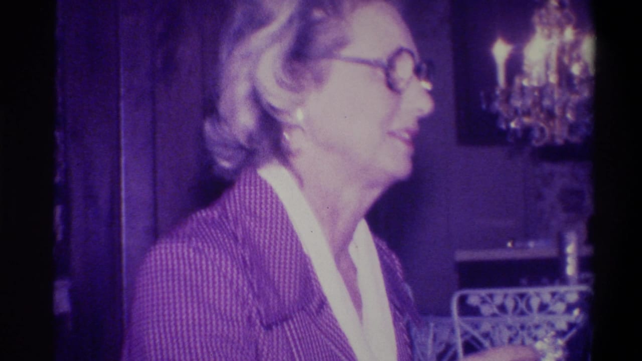Woman in profile wearing patterned jacket and round glasses in a domestic interior; crystal chandelier and ornate decorative furniture visible behind her; Troy, New York, 1973 8mm home movie.