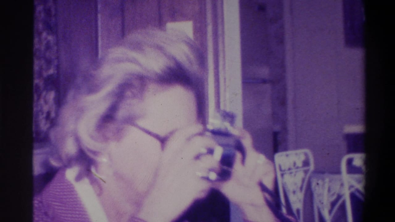 Close-up of woman in round glasses raising a compact still camera to her eye and smiling; window visible in background; Troy, New York, 1973 8mm home movie.