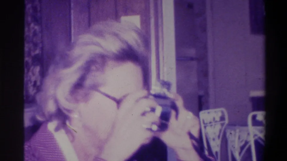 Close-up of woman in round glasses raising a compact still camera to her eye and smiling; window visible in background; Troy, New York, 1973 8mm home movie.