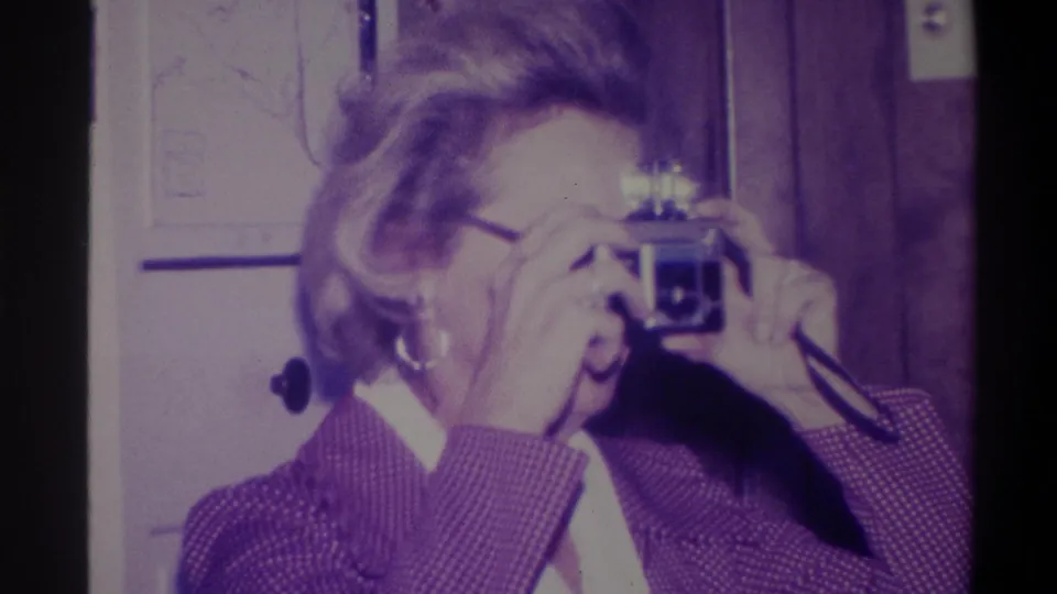 Woman looking through a compact still camera viewfinder in a domestic interior; hoop earring and patterned jacket visible; Troy, New York, 1973 8mm home movie.
