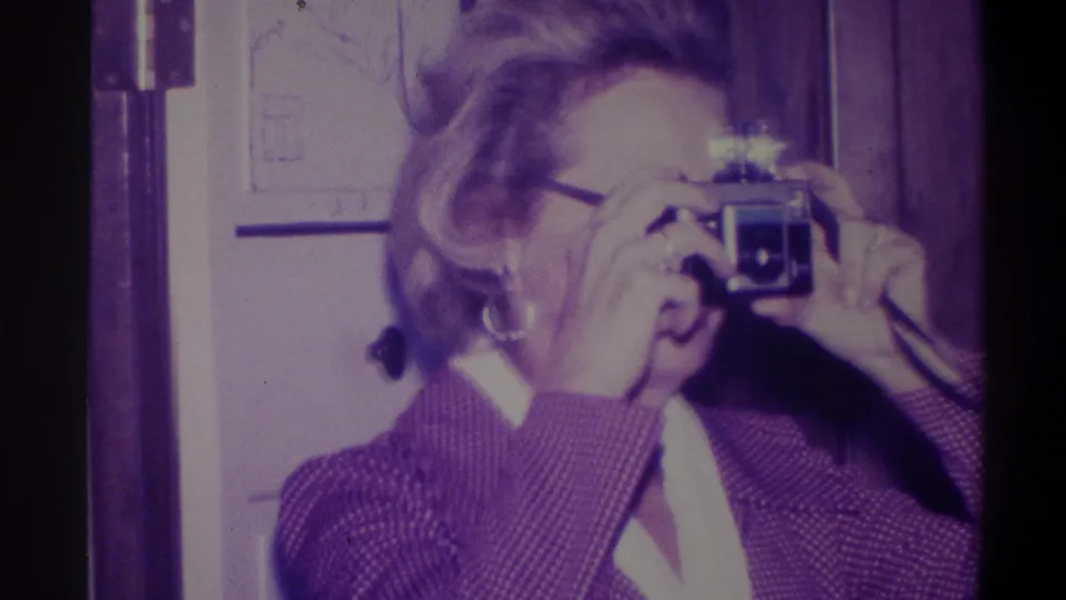 Woman with compact still camera raised to eye, turning slightly toward the viewer; door frame and wall visible in background; 8mm home movie, Troy, New York, 1973.