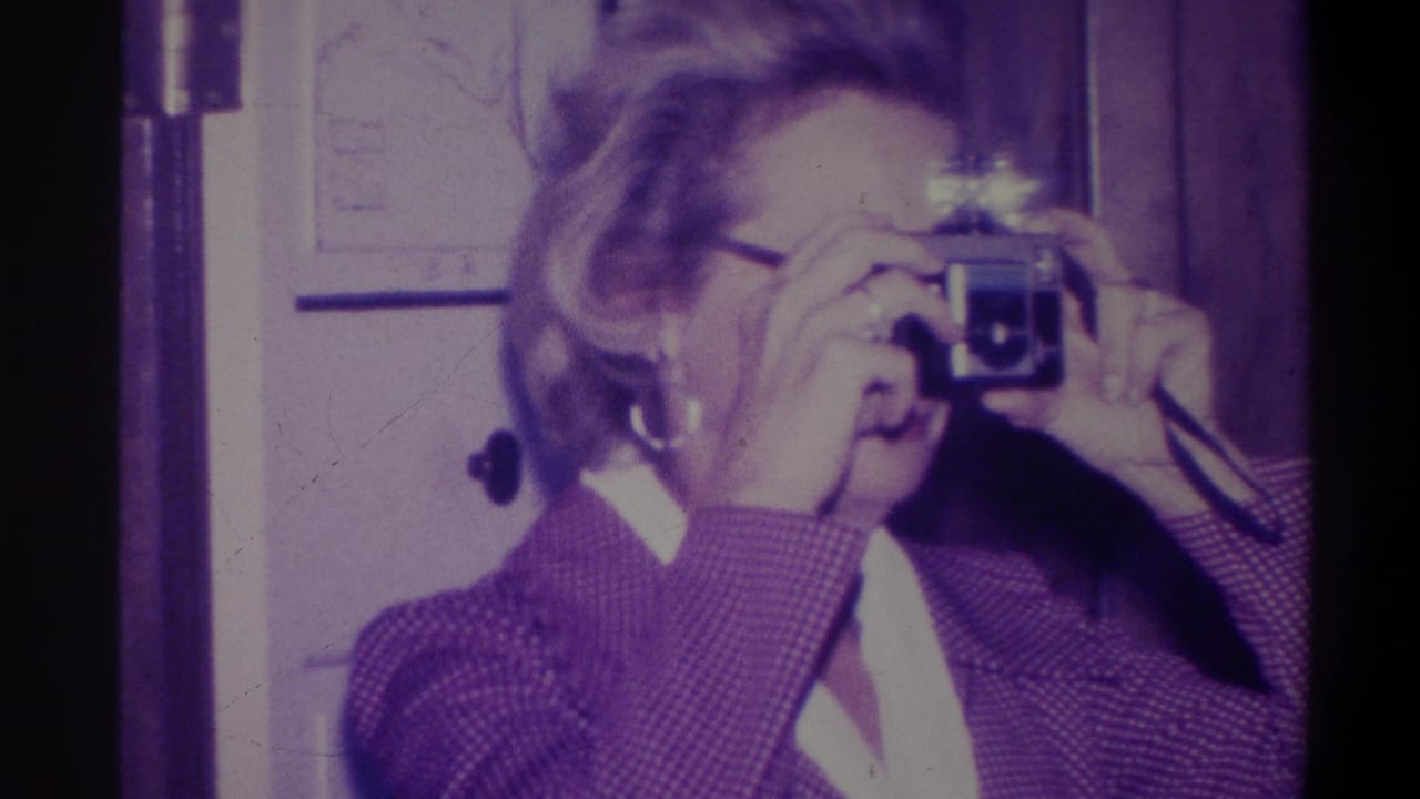 Woman wearing glasses and a purple patterned jacket raises a vintage viewfinder camera; 8mm home movie from Troy, New York, 1973