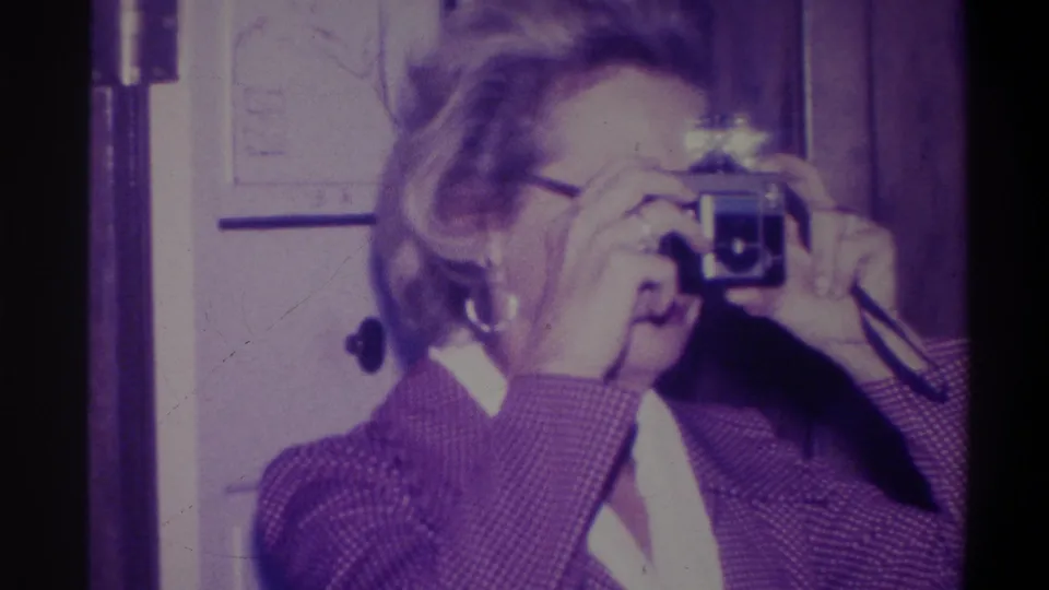 Woman wearing glasses and a purple patterned jacket raises a vintage viewfinder camera; 8mm home movie from Troy, New York, 1973