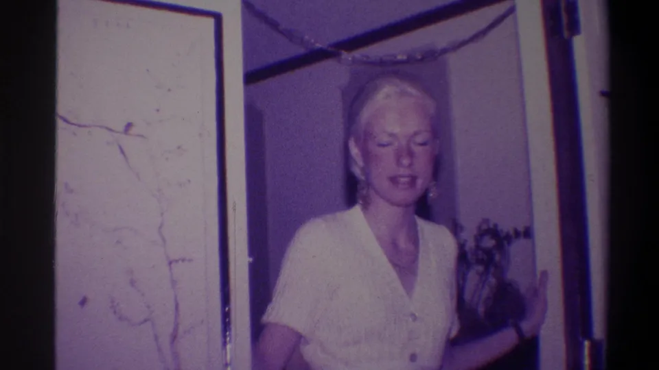 Blonde woman in white top glances downward mid-pose, framed artwork on wall to left; 8mm interior footage from Troy, New York, 1973