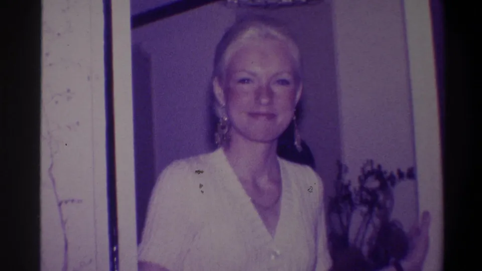 Blonde woman in white short-sleeve top and drop earrings faces the camera directly; door frame and foliage behind her; 8mm home movie, Troy NY, 1973