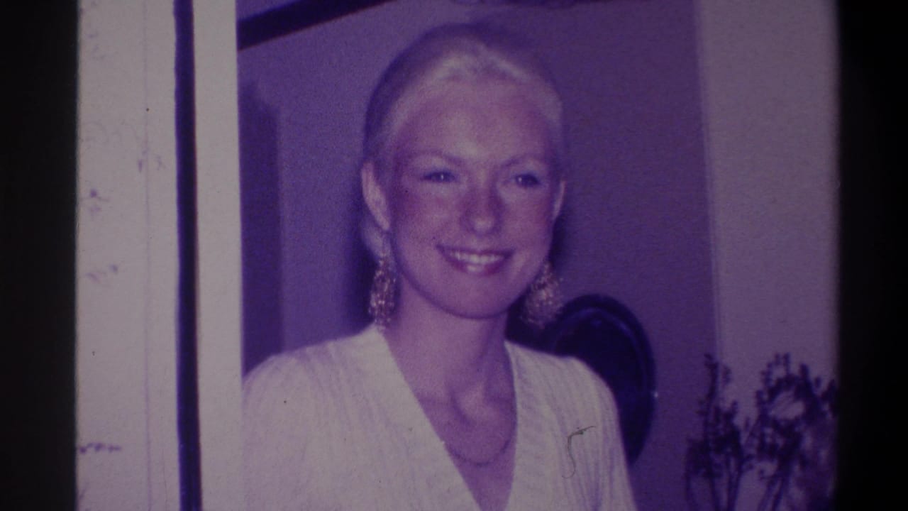 Blonde woman with pulled-back updo smiling at camera, wearing large gold dangling earrings and white V-neck sweater; 8mm home movie, circa 1973, domestic interior, Troy NY.