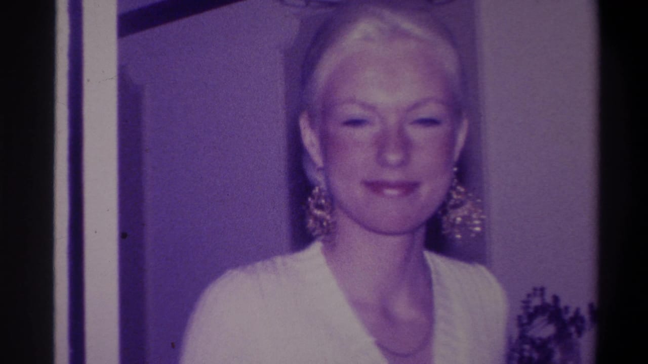 Blonde woman in white V-neck sweater and large gold earrings with composed expression, indoor setting, purple-cast 8mm film.
