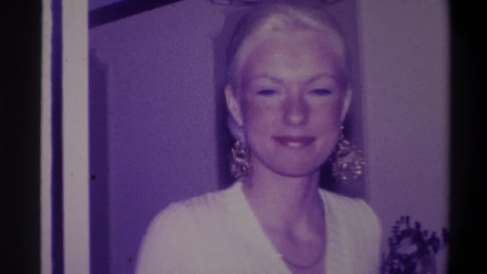 Blonde woman with updo in composed expression, large gold dangling earrings clearly visible, white V-neck sweater; 8mm home movie circa 1973, domestic interior, door frame visible in background.