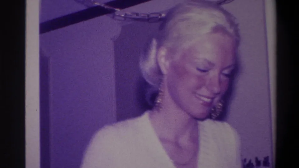 Blonde woman with updo looking downward in a candid moment, gold dangling earrings and white V-neck sweater visible; 8mm home movie circa 1973, domestic interior.