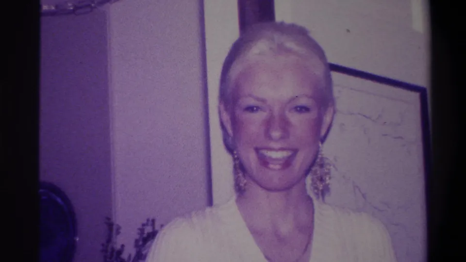 Blonde woman with updo smiling broadly at camera, white V-neck sweater and large gold earrings; domestic interior with framed artwork on wall and flower arrangement visible; 8mm home movie circa 1973.