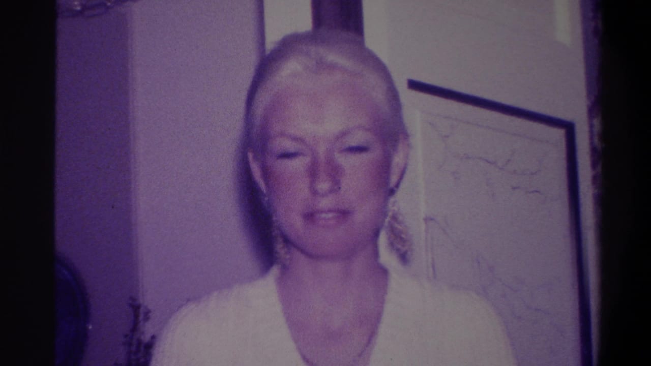 Woman with blonde hair and dangling earrings in white V-neck dress, looking downward in a residential interior; 8mm home movie, Troy, New York, circa 1973.