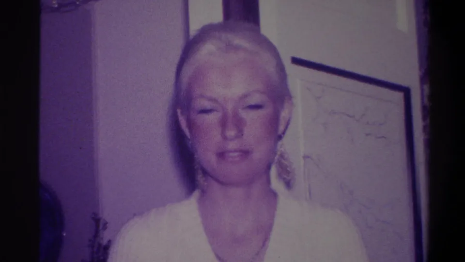 Woman with blonde hair and dangling earrings in white V-neck dress, looking downward in a residential interior; 8mm home movie, Troy, New York, circa 1973.
