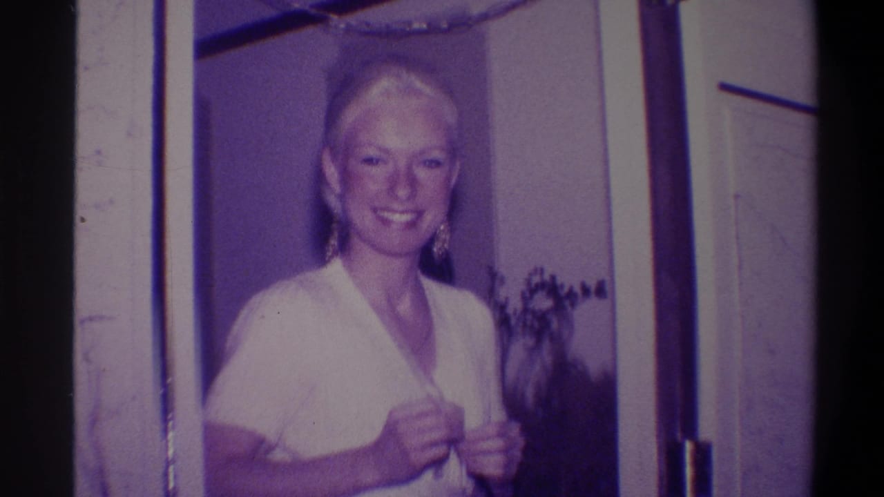Blonde woman smiling broadly in a doorframe, white dress, hands at neckline, houseplant visible behind her.