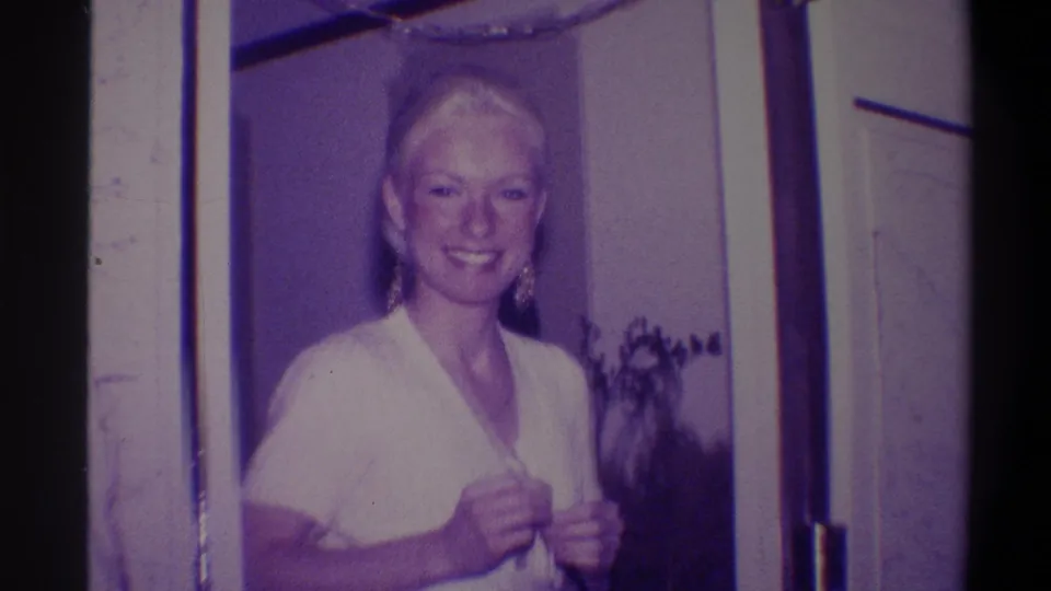 Woman in white V-neck dress smiling warmly in a residential doorframe, hands adjusting neckline; 8mm home movie, Troy, New York, circa 1973.
