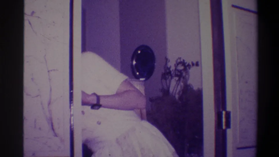 Woman in white dress adjusting her skirt, wristwatch visible on wrist; round wall mirror and houseplant in residential interior; 8mm home movie, circa 1973.
