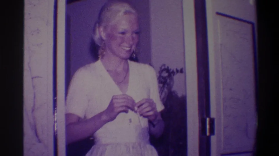 Woman in white V-neck dress smiling and adjusting neckline in a residential doorframe; framed picture and houseplant in background; 8mm home movie, Troy, New York, circa 1973.