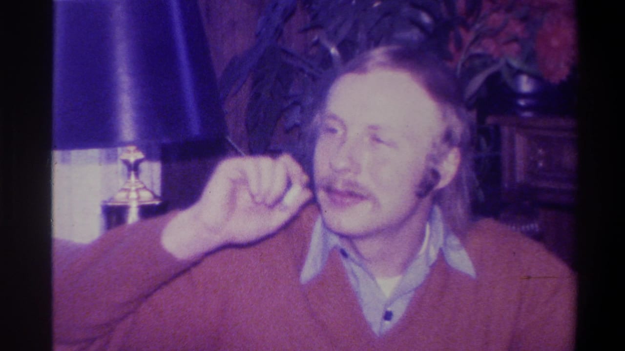 Mustachioed man in red sweater gesturing in a domestic interior, Troy, New York, circa 1973; table lamp and houseplants visible behind him.