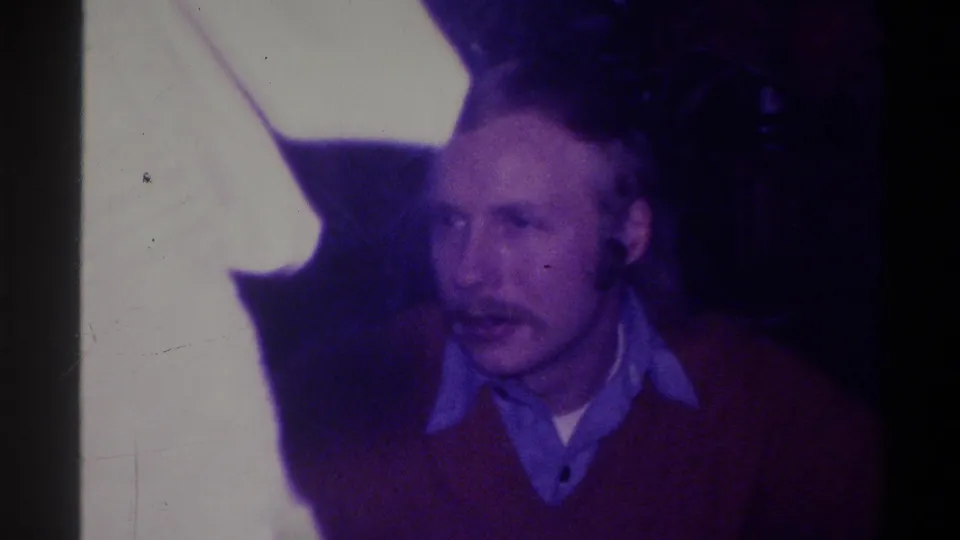 Mustachioed man in red sweater appearing to speak or react in a domestic interior, Troy, New York, circa 1973; blurred foreground element at left.