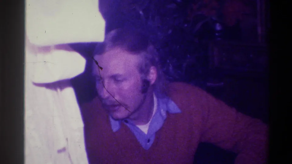 Mustachioed man in red sweater looking downward in domestic interior, Troy, New York, circa 1973; diagonal film scratch artifact visible across frame.