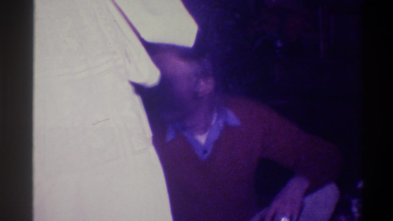 Man in rust sweater leaning forward in a private home interior; strong age-related color shift consistent with 1973 Troy, New York 8mm home movie.