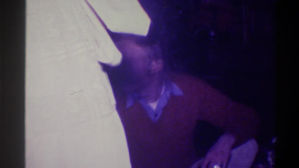 Man in rust sweater leaning forward in a private home interior; strong age-related color shift consistent with 1973 Troy, New York 8mm home movie.
