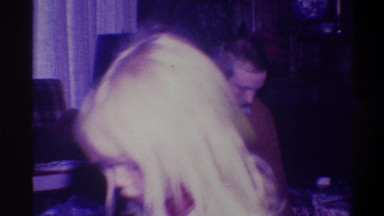 Blonde figure bending forward; second darker-haired person visible behind them in a dim home interior with dark furniture and curtains.