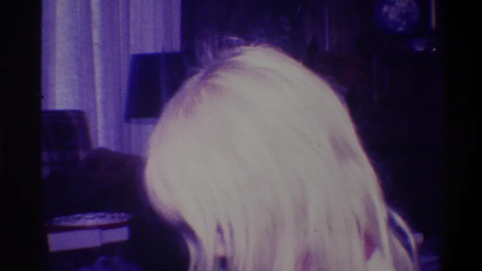 Close rear view of a blonde figure in a 1970s domestic interior; dark sofa and circular wall decoration visible behind; blue-shifted 8mm exposure.
