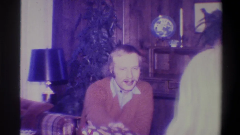 Smiling man in orange-rust cardigan with a colorful wrapped gift in a wood-paneled living room, consistent with a 1973 Troy, New York Christmas home movie.