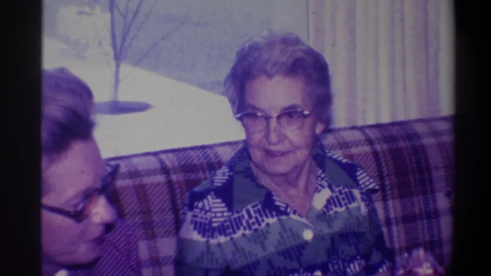 Elderly woman in geometric patterned blouse on plaid couch, facing camera; second person at frame left; bare winter tree through window behind her, Troy, New York, circa 1973.