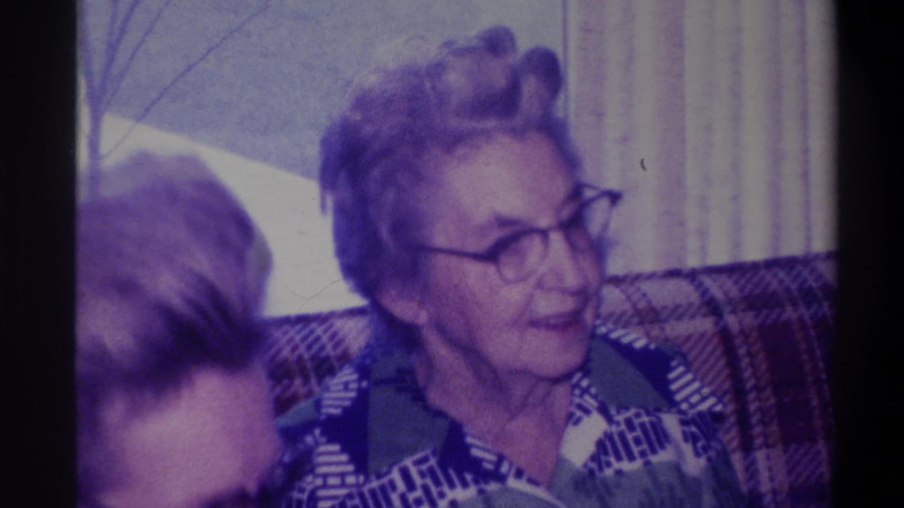 Elderly woman with glasses mid-expression, mouth slightly open; second person's dark hair visible at left; plaid couch and curtained window behind her.
