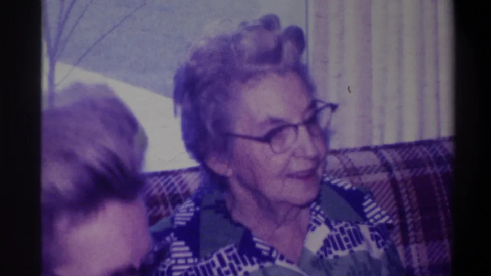 Elderly woman in 1970s patterned blouse speaking or reacting on plaid couch; companion's hair at frame left; curtained window and bare tree behind her, circa 1973.