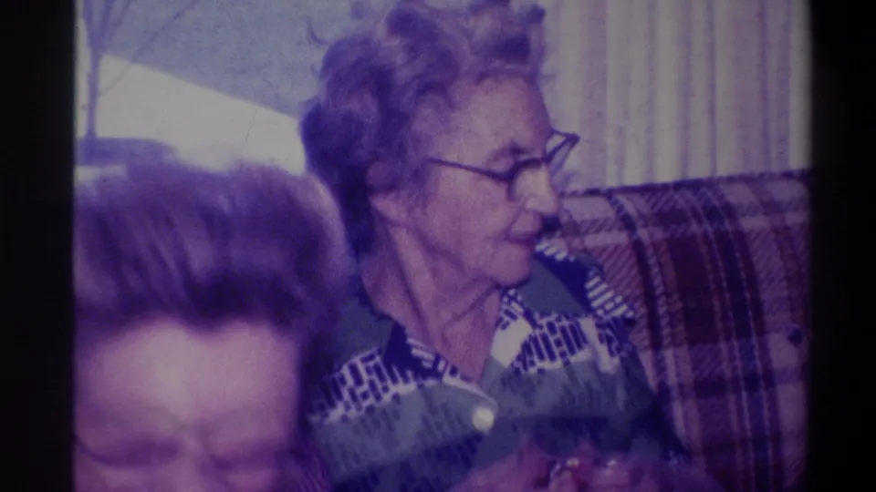 Elderly woman in three-quarter profile on plaid couch engaging with a companion at frame edge; bare winter tree through window, Troy, New York, circa 1973, 8mm home movie.