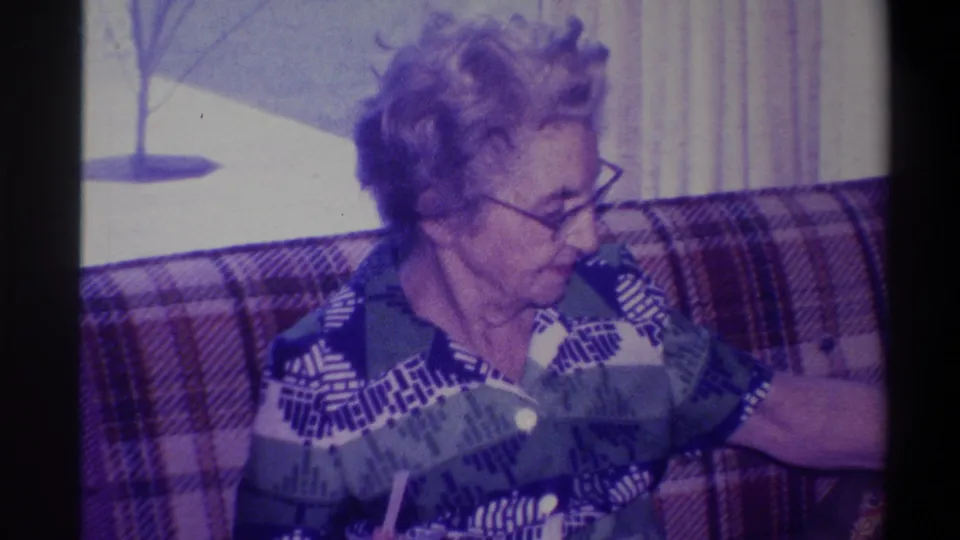 Elderly woman on plaid couch in three-quarter profile with animated expression during family conversation; bare winter tree through window, circa 1973, 8mm home movie.