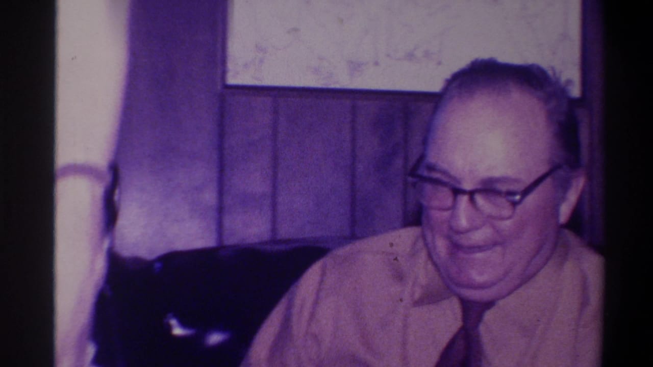 Older man in thick-framed glasses and light shirt in a 1970s wood-paneled domestic interior; 8mm footage attributed to Troy, New York, 1973.