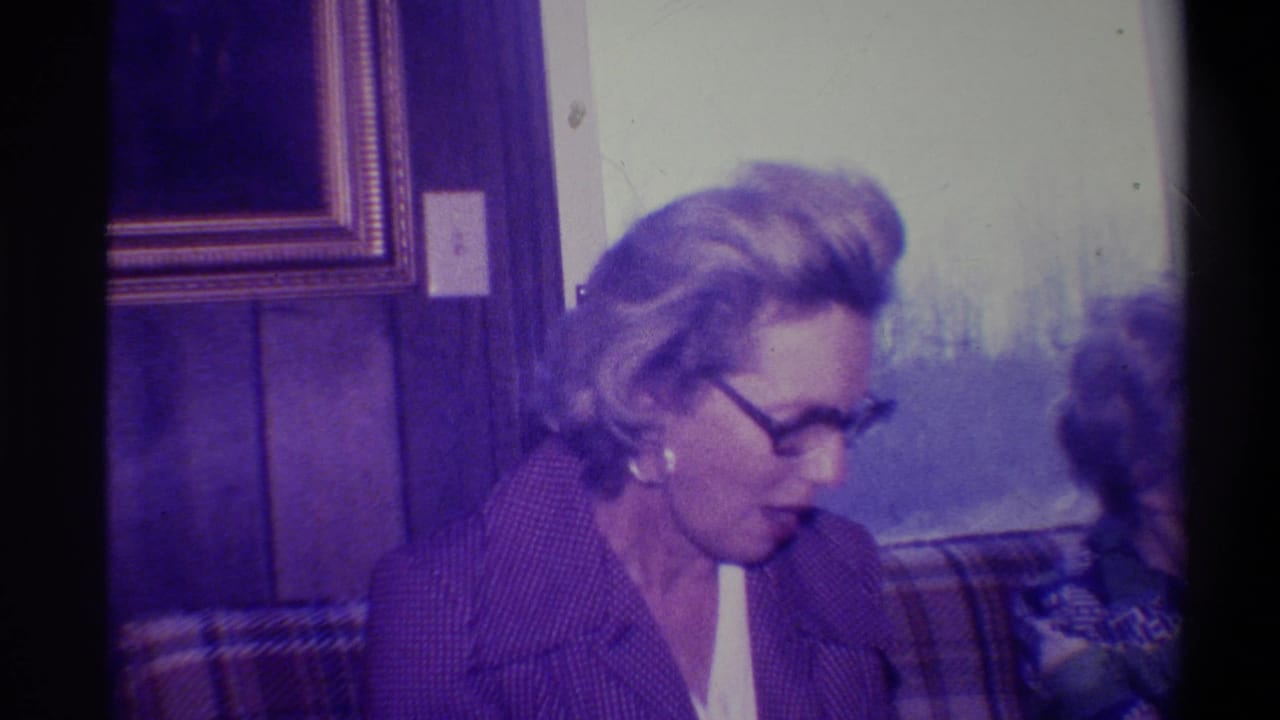 Woman with light hair and glasses in a dark patterned jacket seated near wood-paneled wall; gold-framed picture and plaid upholstery visible.