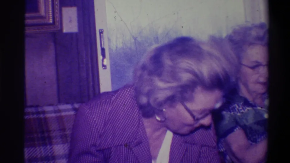 Woman with glasses leaning forward in apparent laughter, second older woman visible behind her in a 1970s wood-paneled domestic interior; 8mm footage, Troy, New York, 1973.