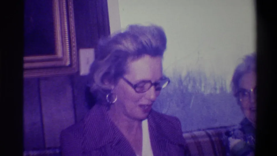 Woman in dark patterned jacket with hoop earrings and glasses in a 1970s wood-paneled domestic interior; 8mm footage attributed to Troy, New York, 1973.