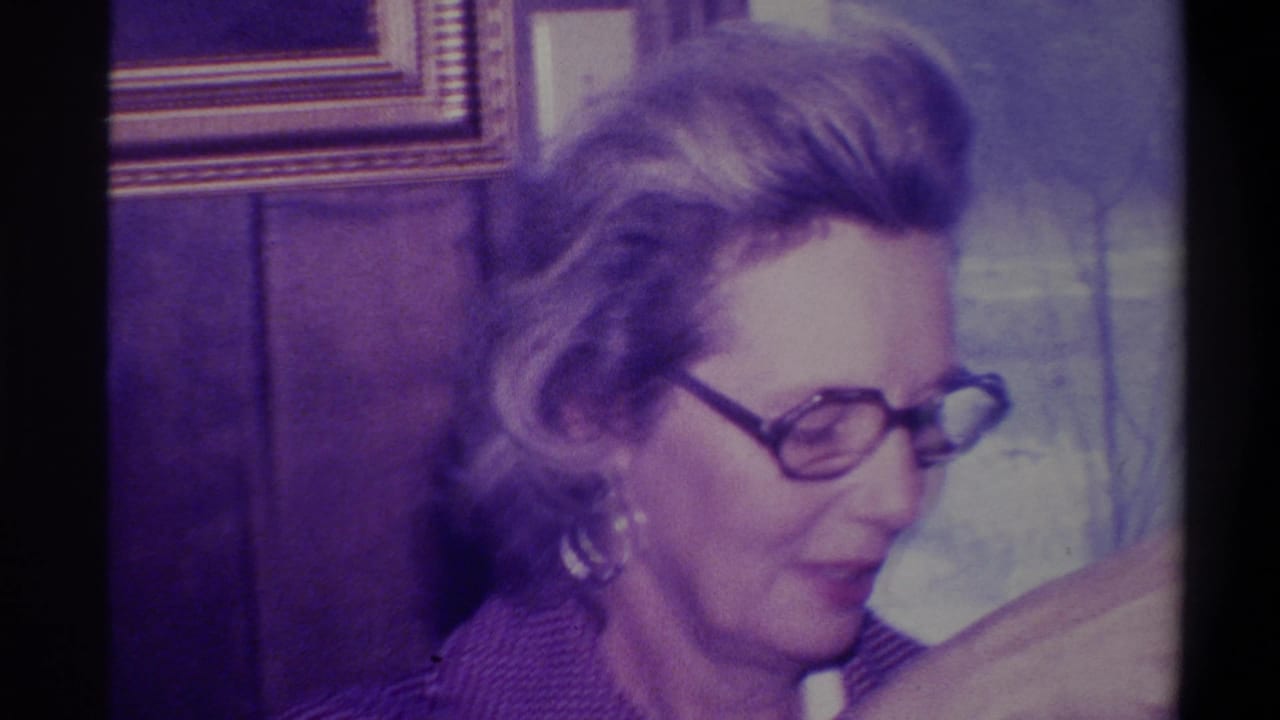 Woman in profile wearing dark-rimmed glasses against a wood-paneled wall with a gilt-framed picture.