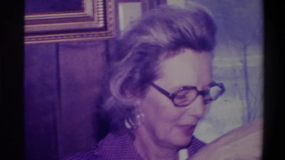 Woman in profile with styled blonde hair and dark-rimmed glasses; wood-paneled interior with gilt-framed picture, 8mm footage circa 1973, Troy, New York.