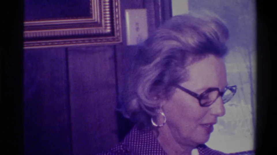 Woman with styled blonde hair, dark-rimmed glasses, and large hoop earring in a wood-paneled domestic interior; 8mm footage circa 1973, Troy, New York.