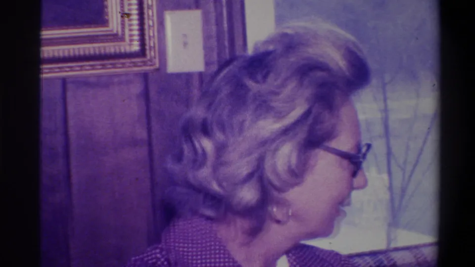 Woman with styled wavy hair and partial glasses visible, turned from camera in a wood-paneled interior; 8mm footage circa 1973, Troy, New York.