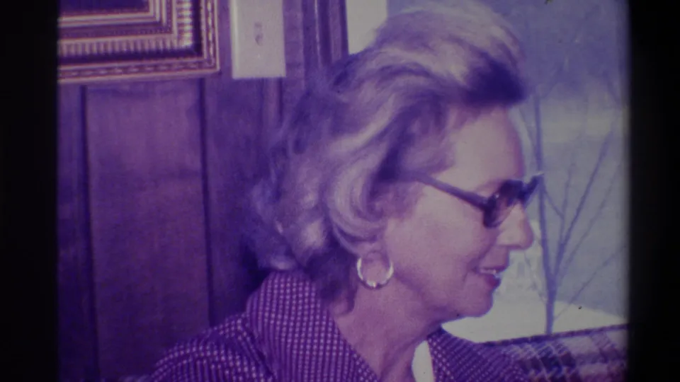 Profile view of a woman with styled blonde bouffant hair, dark-rimmed glasses, and gold hoop earring in a wood-paneled domestic interior, Troy, New York, circa 1973