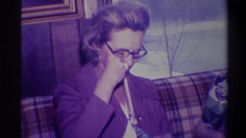 Woman with dark-rimmed glasses and gold hoop earring turning toward camera with hand raised, plaid upholstered sofa visible in background, Troy, New York, circa 1973
