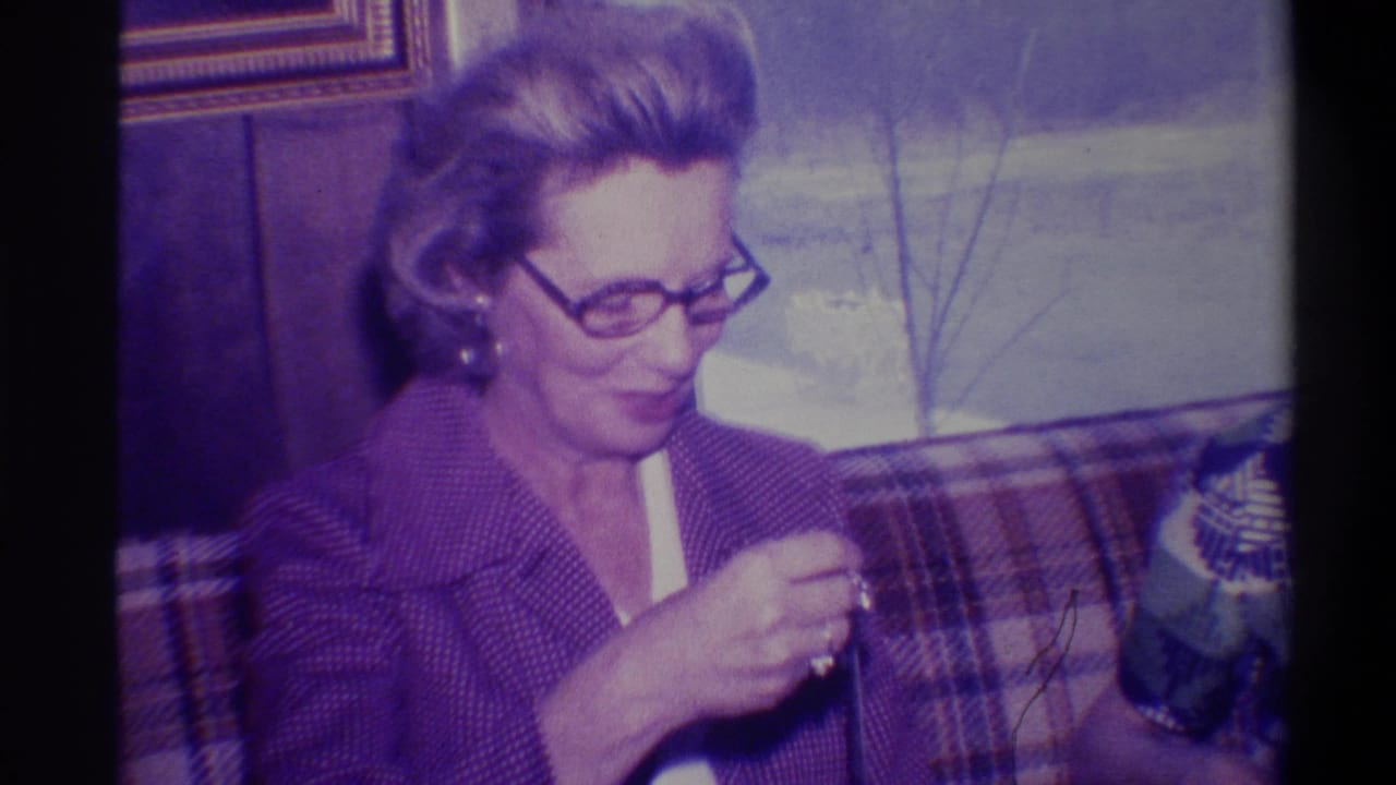 Middle-aged woman in 1970s-style glasses and purple knit jacket opens a gift on a plaid couch; wood-paneled wall and gold-framed picture behind her, Troy, New York, circa 1973.