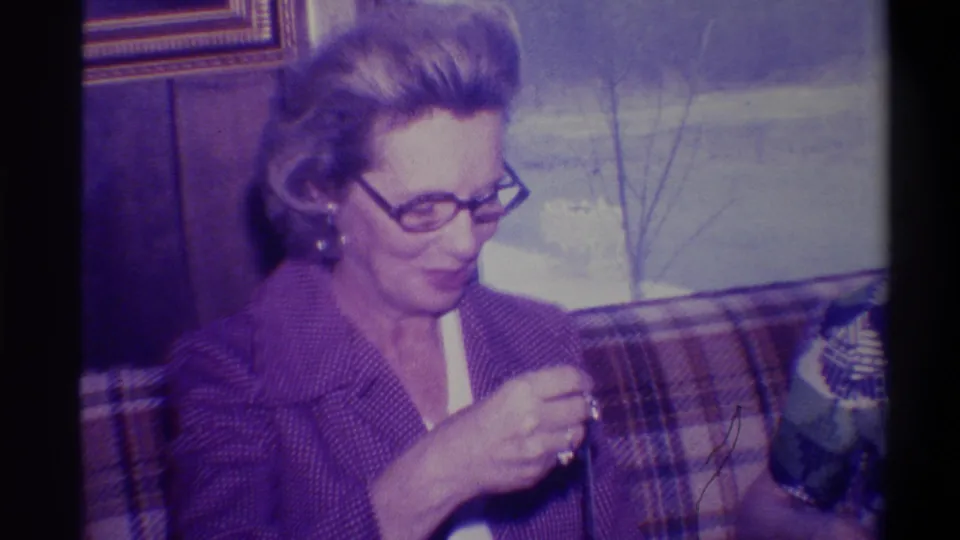Middle-aged woman in 1970s-style glasses and purple knit jacket opens a gift on a plaid couch; wood-paneled wall and gold-framed picture behind her, Troy, New York, circa 1973.