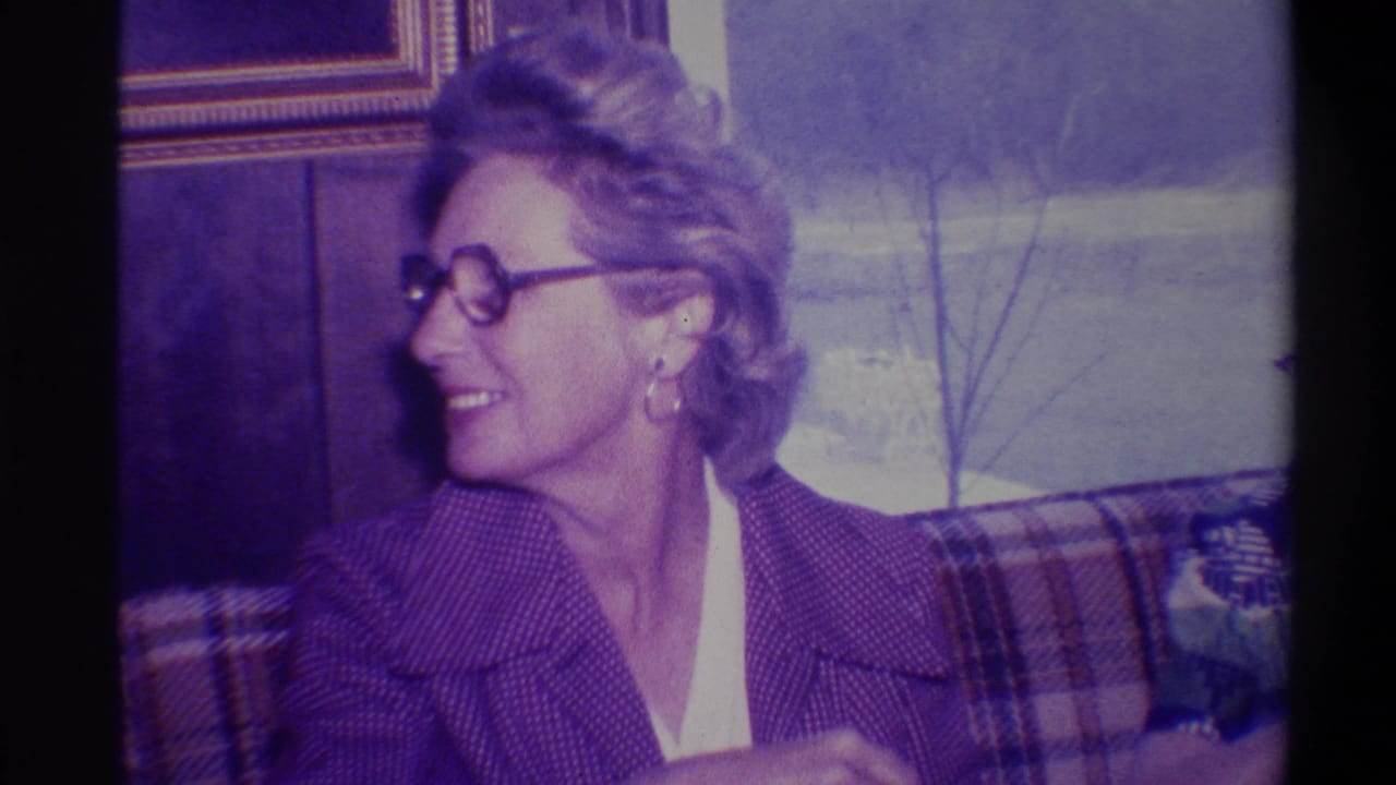 Woman in 1970s-style glasses and purple knit jacket smiles and turns away from camera; plaid couch and wrapped gift partially visible, Troy, New York, circa 1973.