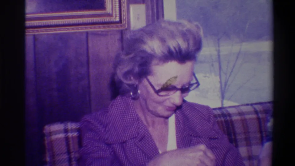 Woman in 1970s-style glasses and purple knit jacket looks down smiling at an opened gift; wood-paneled domestic interior, Troy, New York, circa 1973.