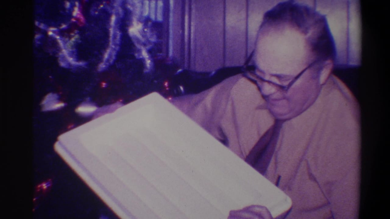 Older man in light button-up shirt and glasses examines a flat white rectangular object in a wood-paneled room, lit Christmas tree behind, 1973 Troy New York home movie.