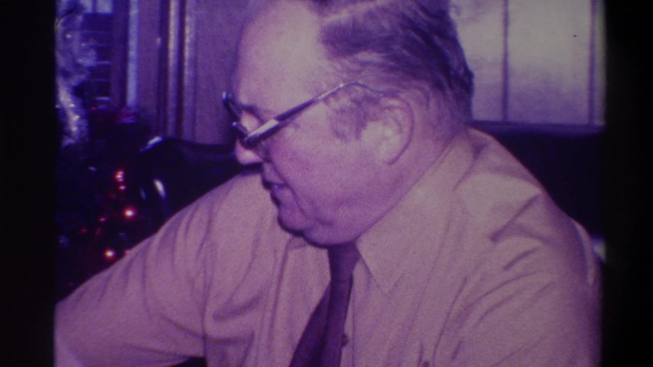 Side profile of older man wearing heavy-framed glasses and a light shirt with dark tie, Christmas tree with red lights behind, 1973 Troy New York home movie.