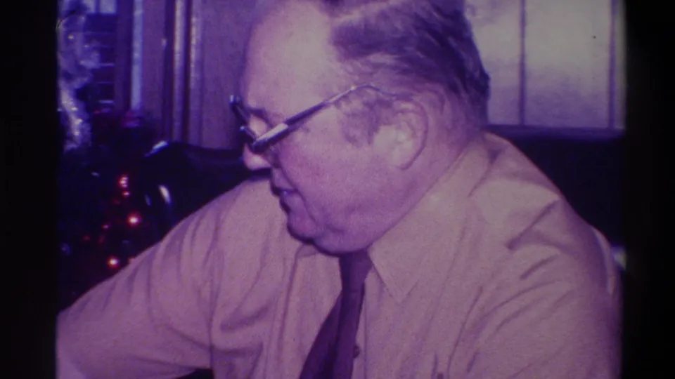 Side profile of older man wearing heavy-framed glasses and a light shirt with dark tie, Christmas tree with red lights behind, 1973 Troy New York home movie.